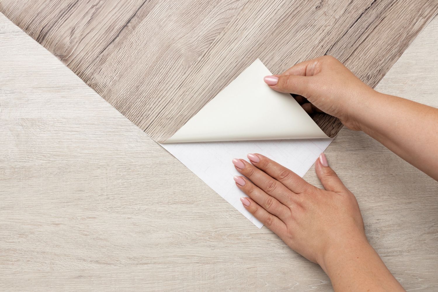 Hands peeling off the backing of a self-adhesive vinyl plank on a light wood floor.
