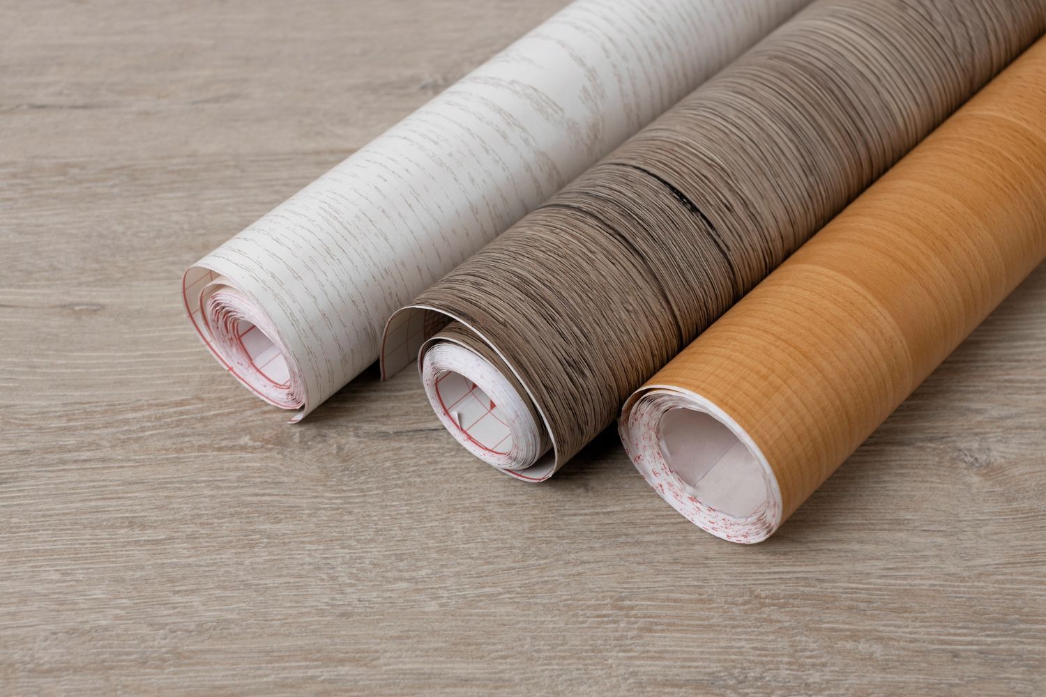 Three rolls of wood-grain contact paper in light beige, brown, and tan, lying on a wooden surface.
