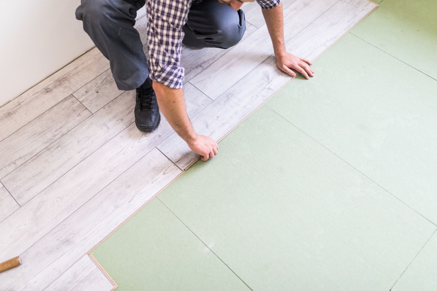 Person kneeling, installing light green flooring over existing wood flooring, indoors.