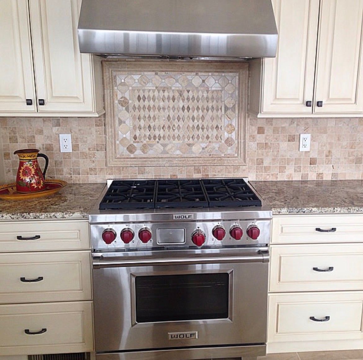 Stainless steel oven with red knobs, beneath a range hood and beige cabinets in a kitchen.