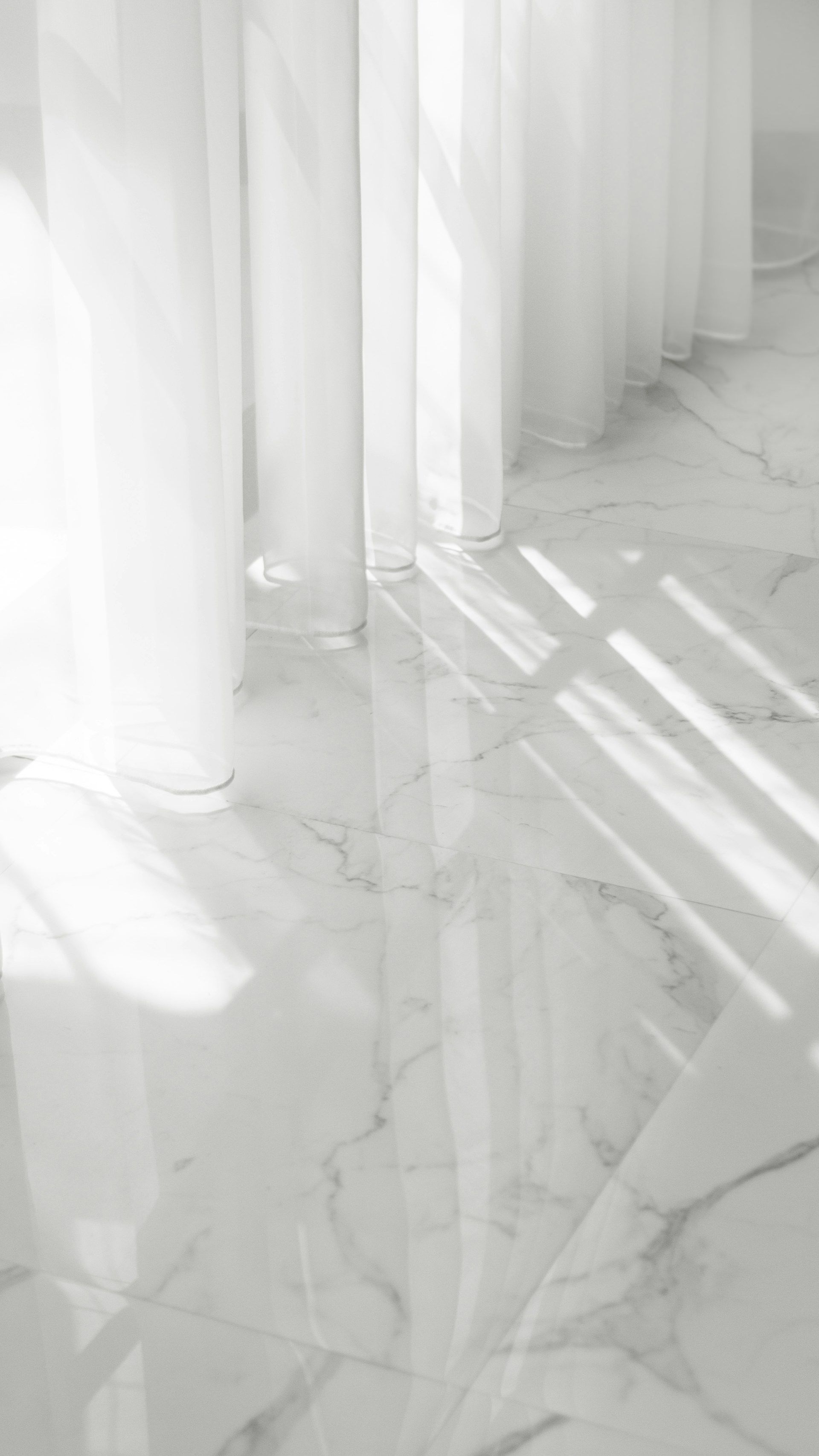 White sheer curtains cast shadows on a white marble floor.