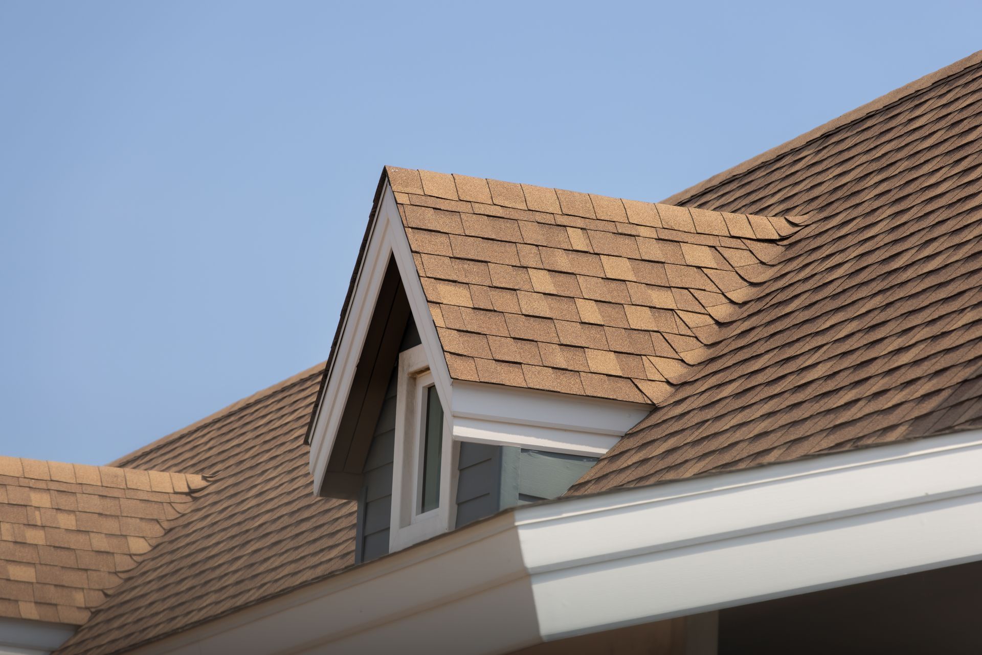 Brown shingled roof with a dormer window against a blue sky.
