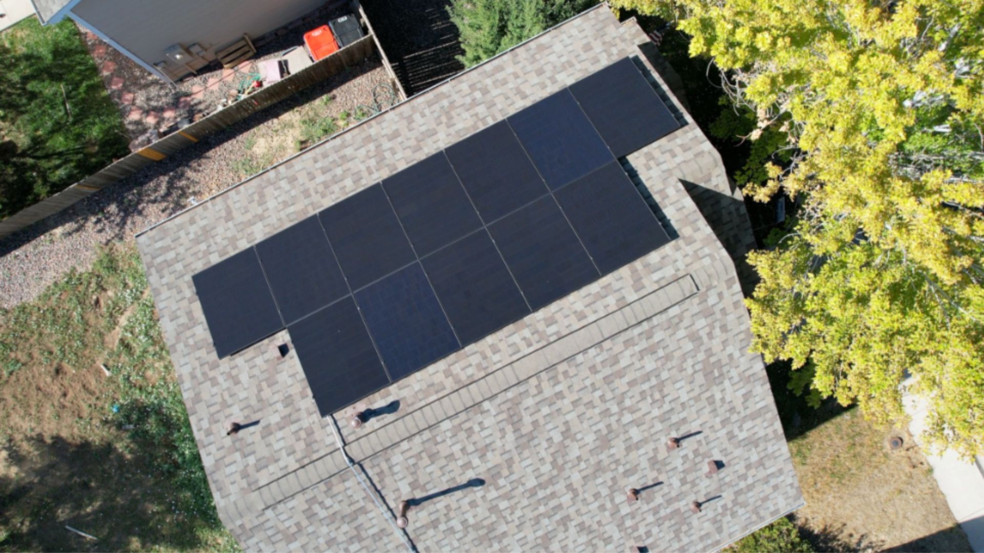 Solar panels on a gray shingled roof, viewed from above, surrounded by green trees and grass.
