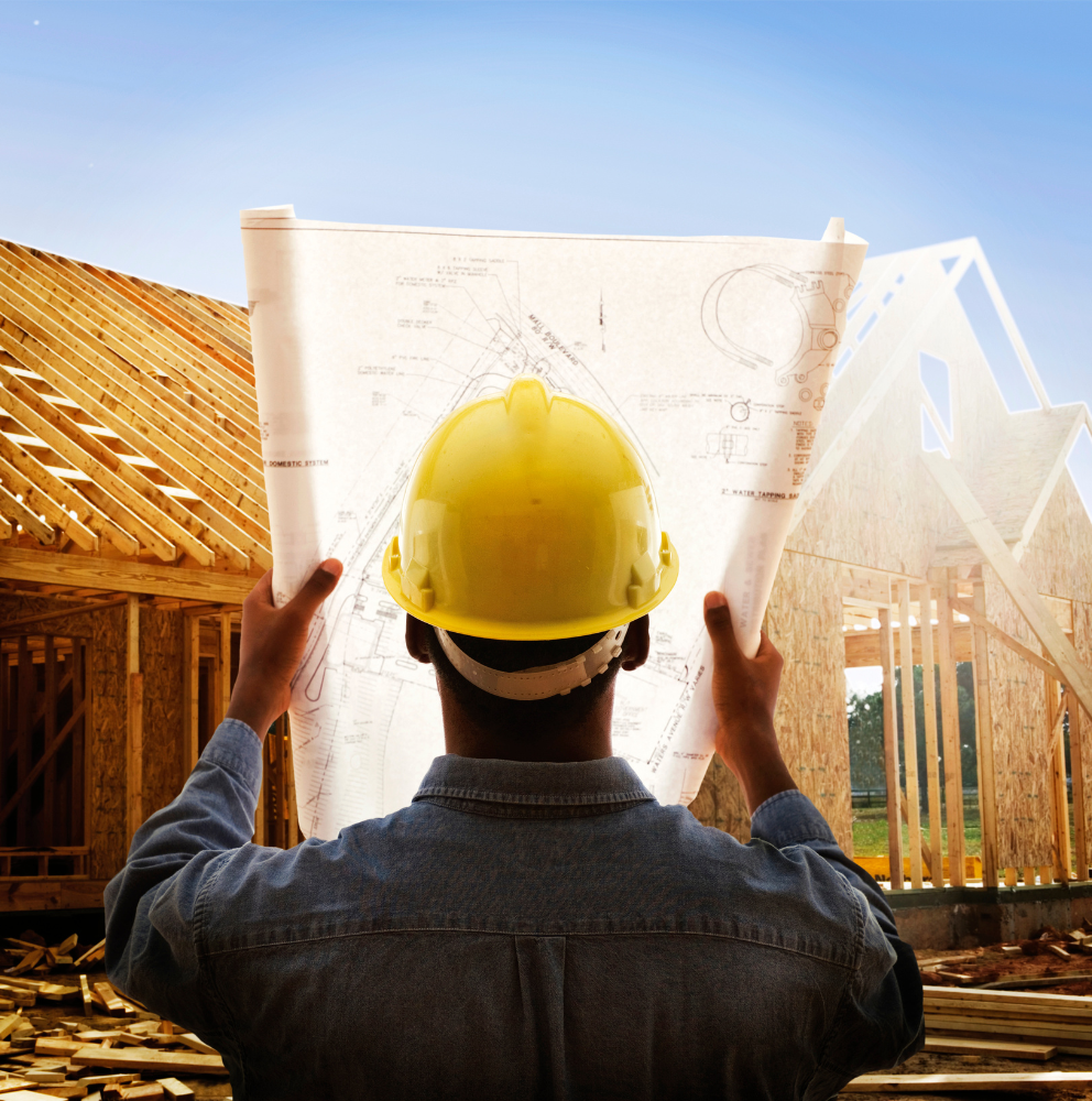 A man wearing a hard hat is holding a blueprint in front of a house under construction