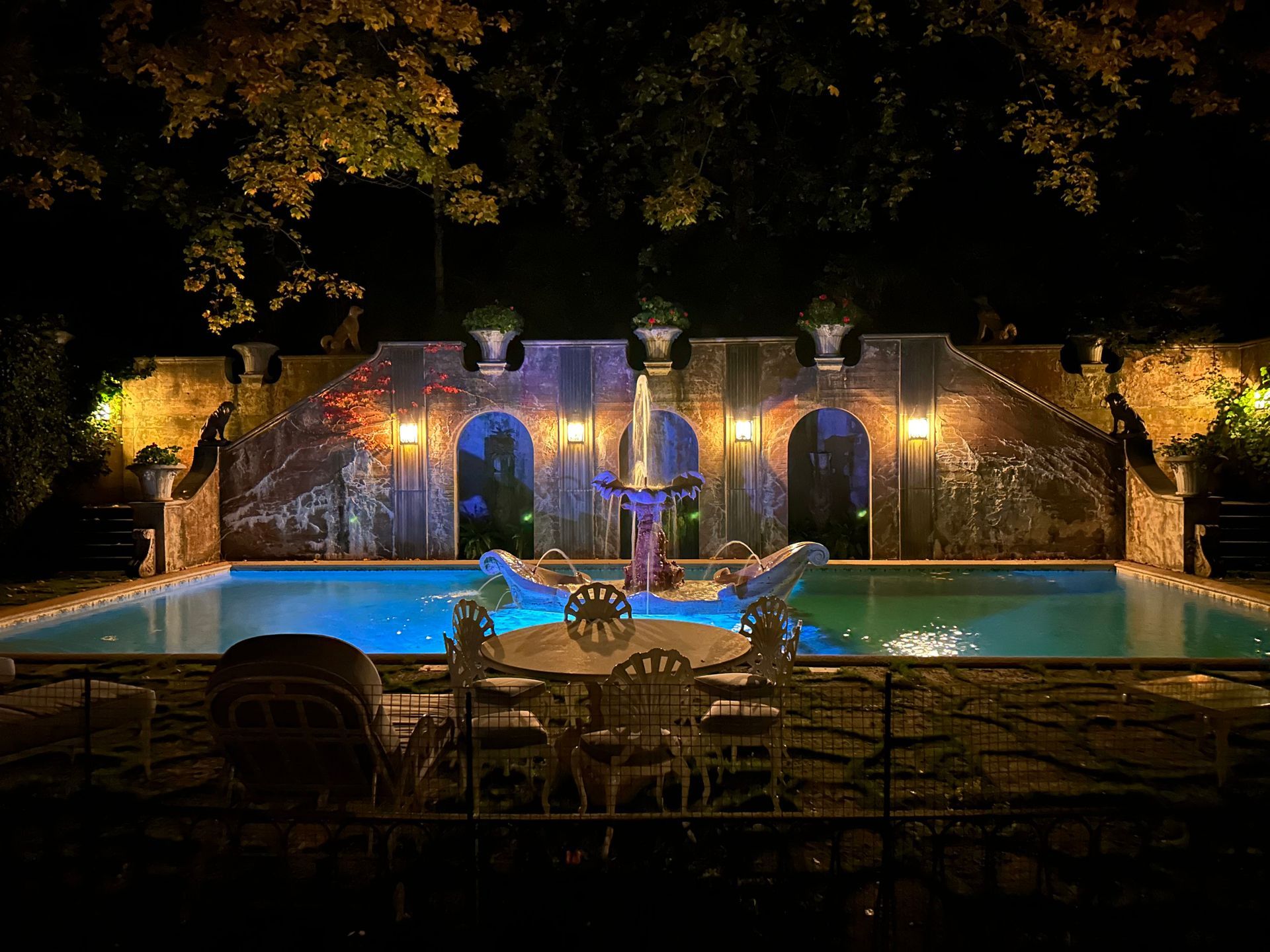 A large swimming pool is lit up at night with a table and chairs in front of it.