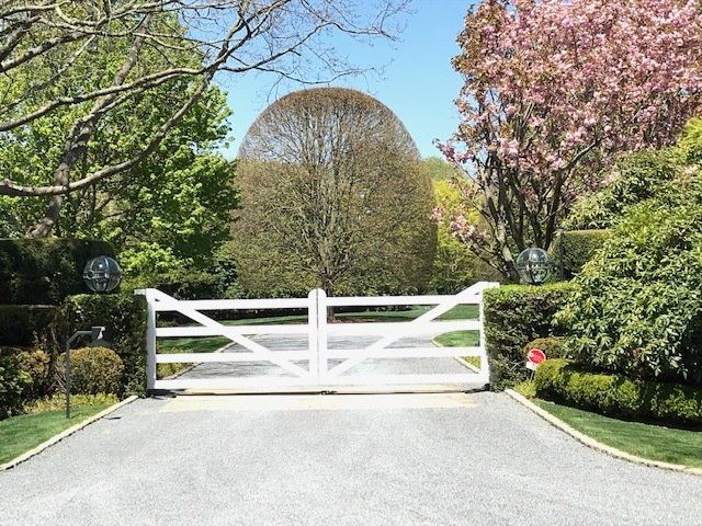 A white gate is in the middle of a driveway surrounded by trees and bushes.
