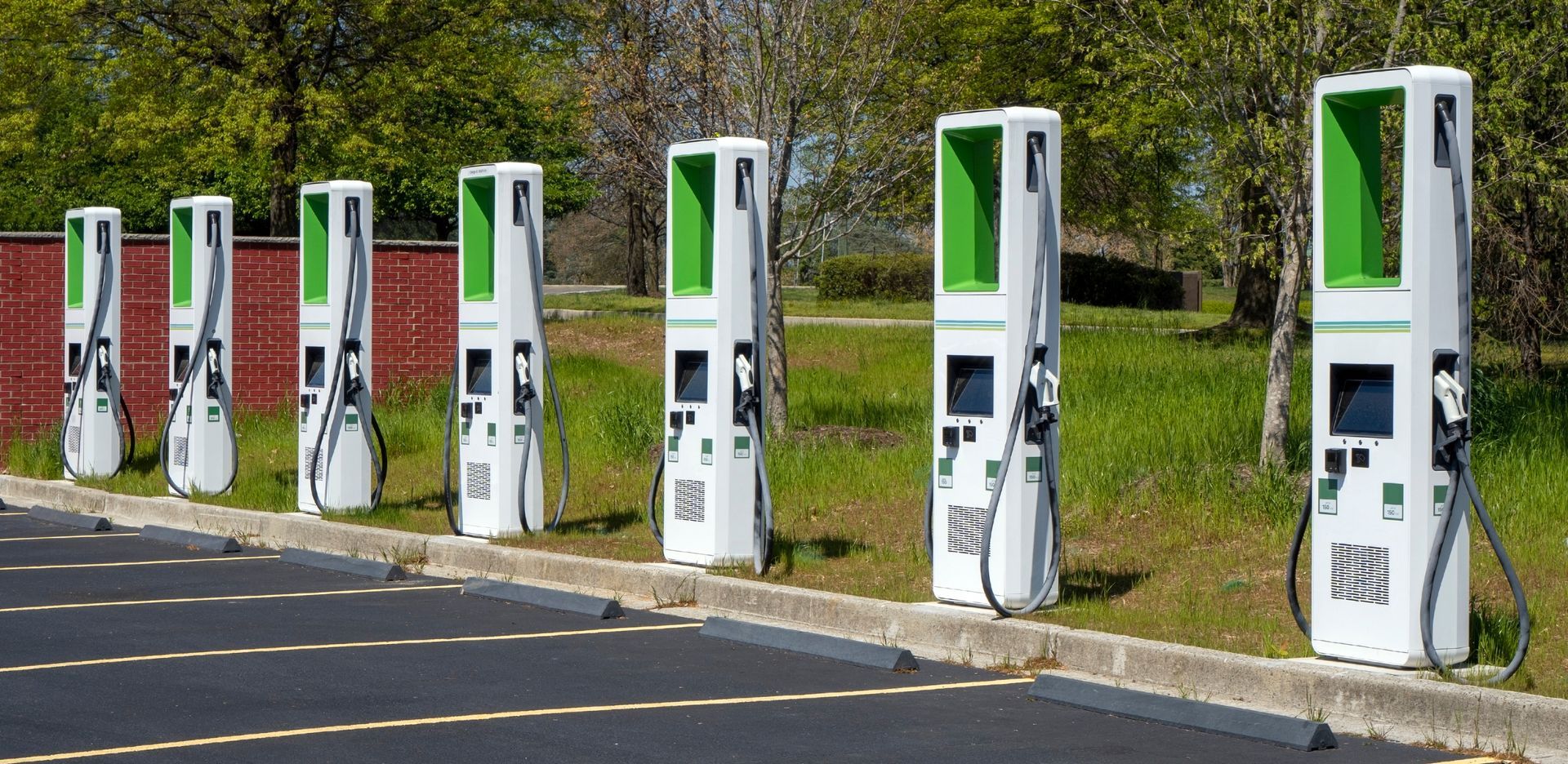 Une rangée de bornes de recharge pour véhicules électriques, blanches avec des touches de vert, se dresse à côté d'un parking. Un feuillage luxuriant apparaît en arrière-plan.