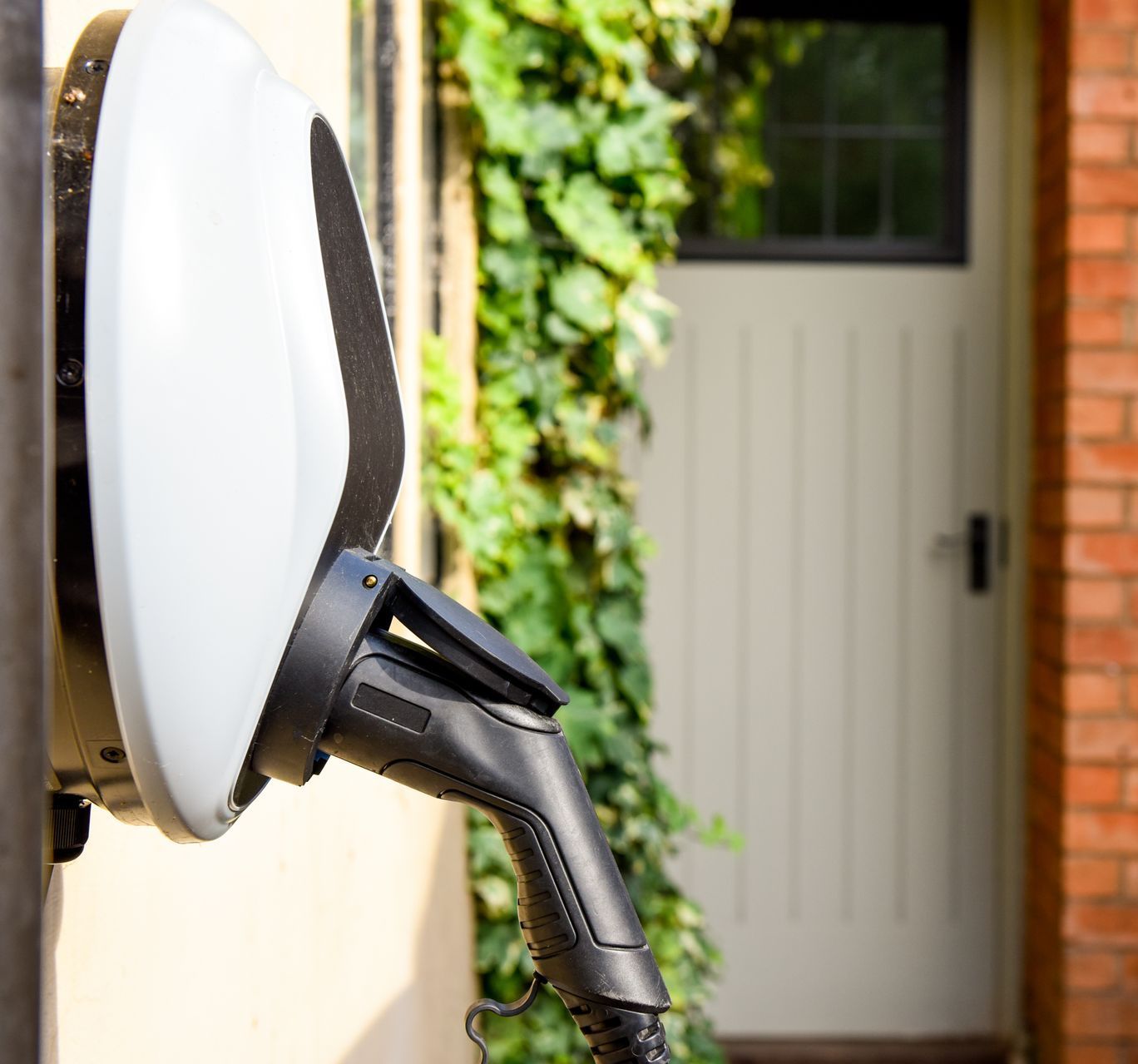 Chargeur blanc pour véhicule électrique fixé sur un mur beige, près d'une porte bordée de verdure. Le câble de charge pend.