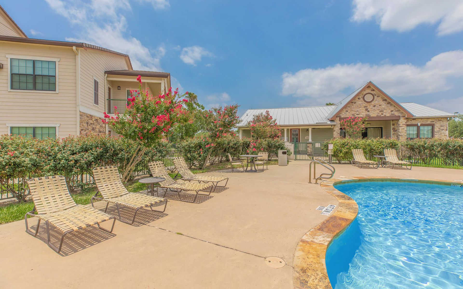 Pool area with lounge chairs, blooming bushes, and a building under a blue sky.