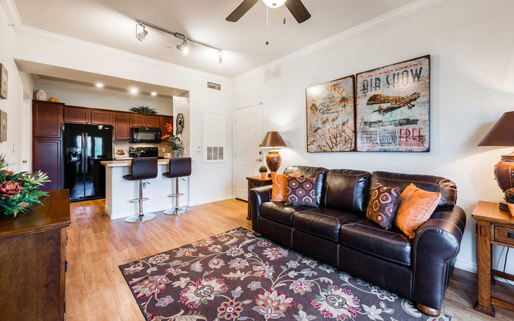 Living room with brown leather sofa, rug, open kitchen with bar stools, and artwork.