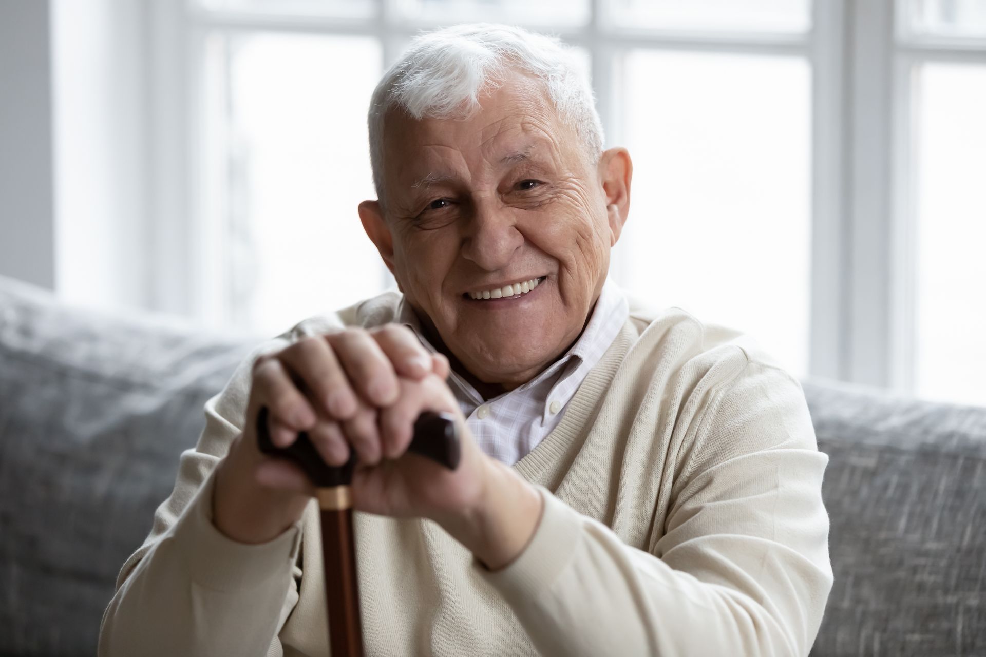 An elderly man is sitting on a couch holding a cane and smiling.