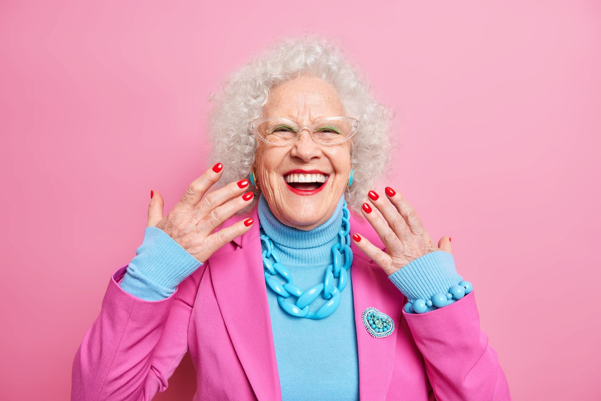 An elderly woman wearing a pink jacket and blue sweater is laughing.