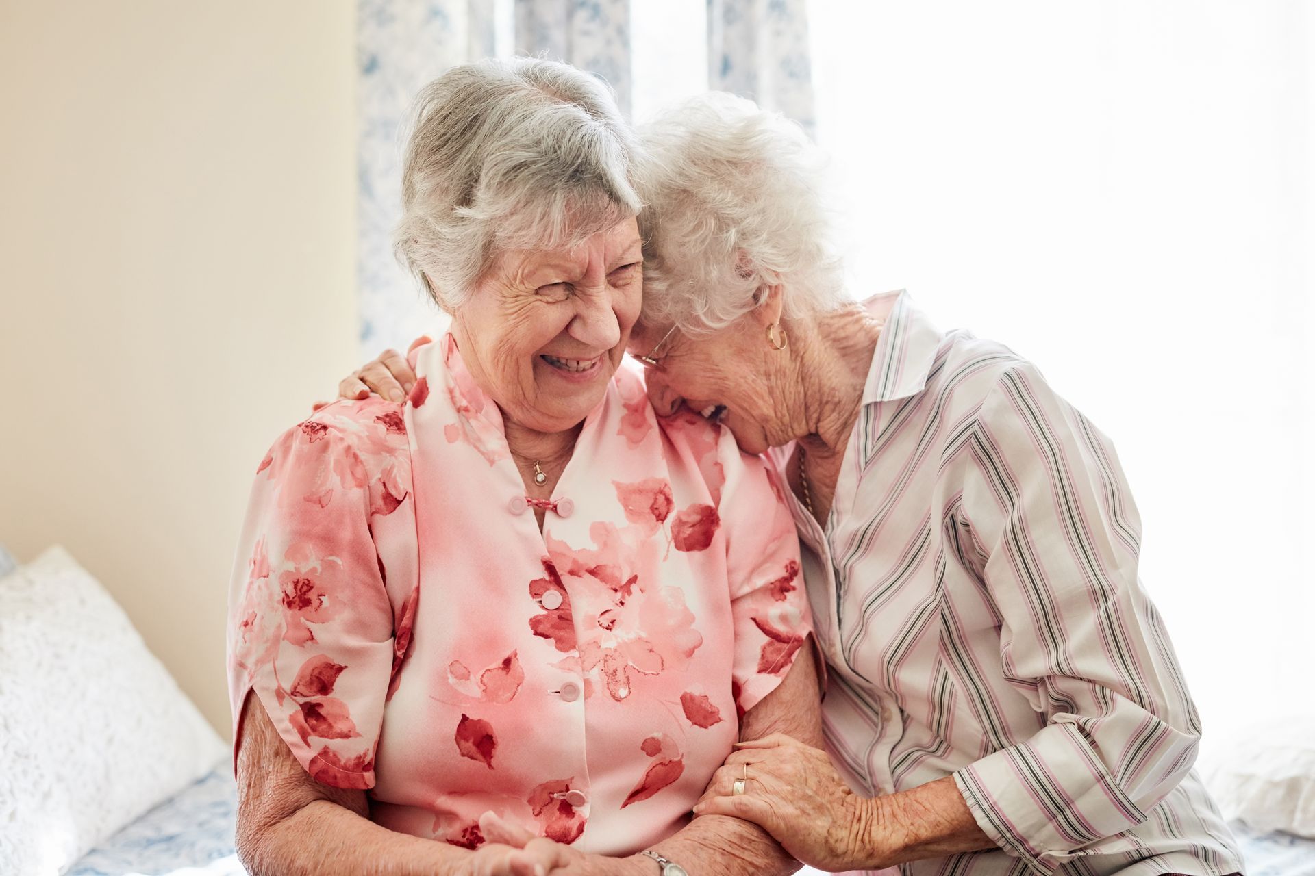 Two elderly women are sitting on a bed holding hands and smiling.