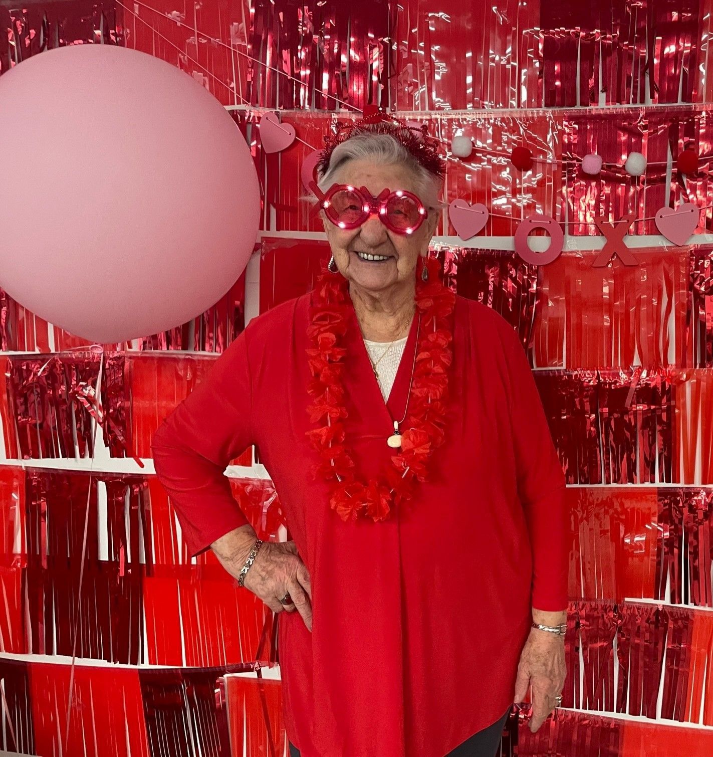 Woman in red outfit and heart-shaped glasses poses in front of a red backdrop with a pink balloon.