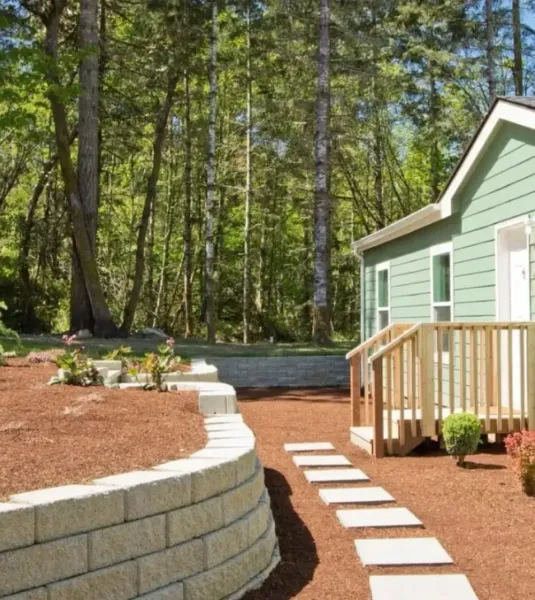 A green house with a wooden porch and stone pathway surrounded by forest.
