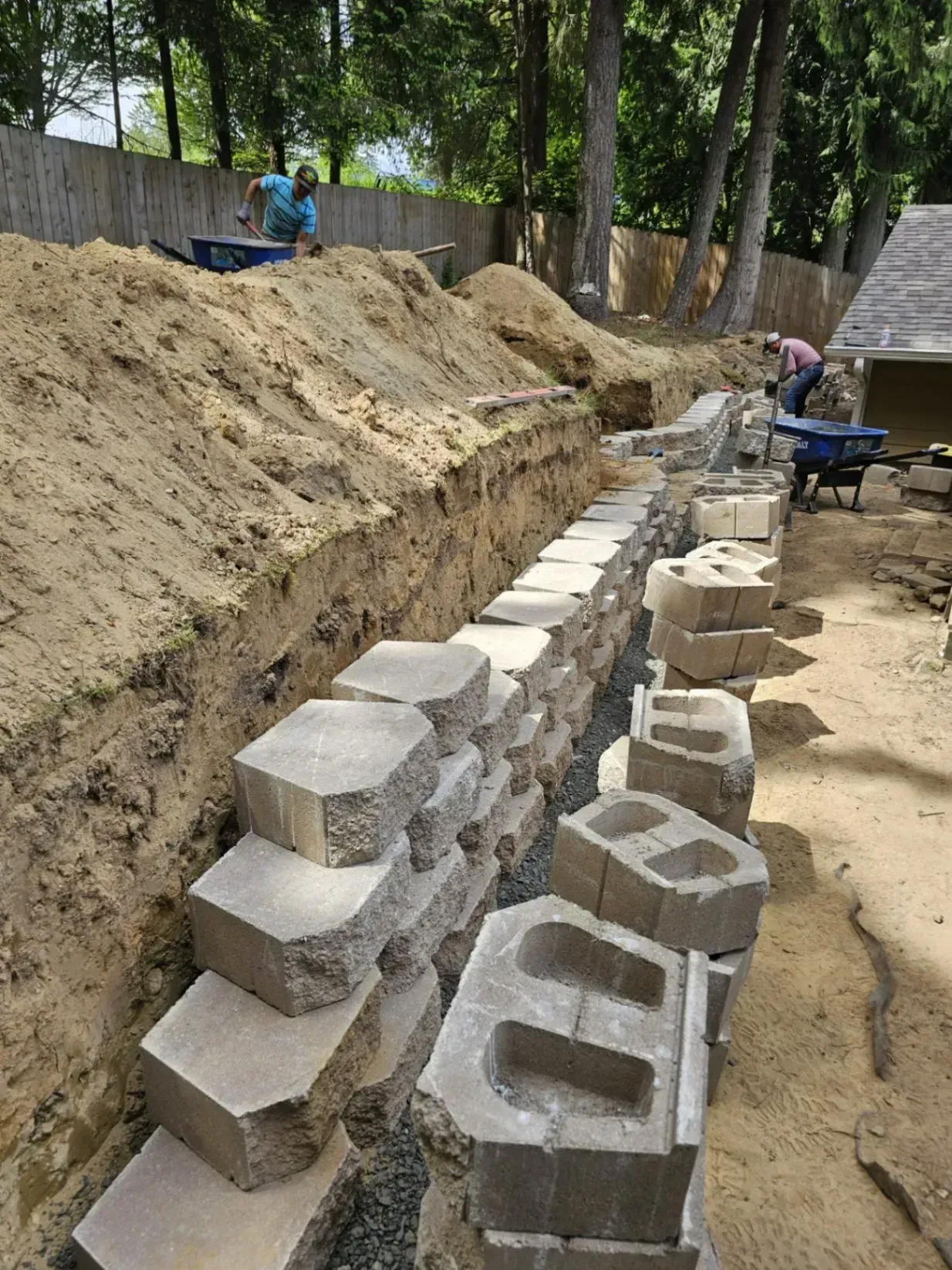 Construction of a retaining wall with concrete blocks. Workers fill and stack blocks in an outdoor setting.