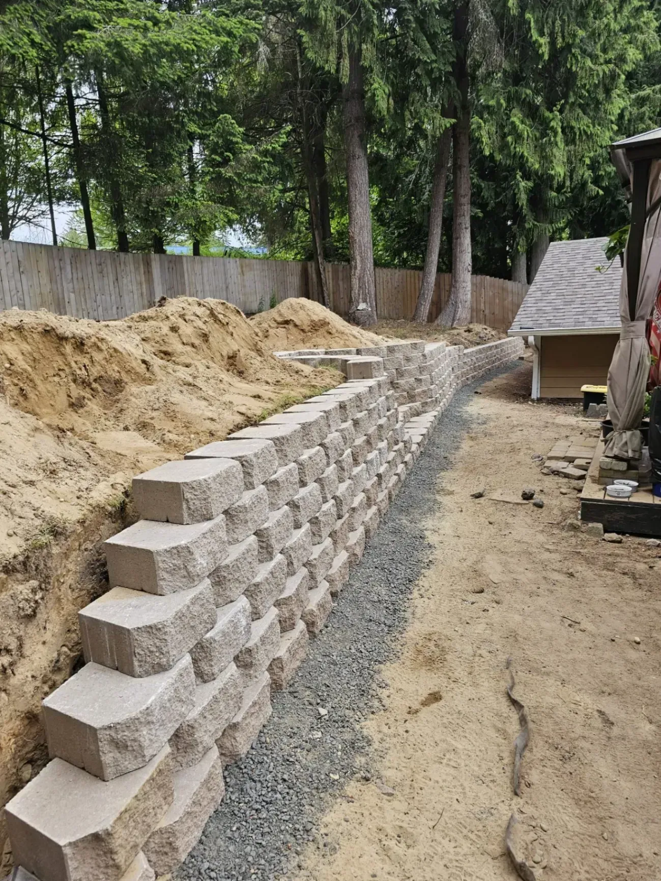 A retaining wall made of stacked concrete blocks next to gravel pathway, leading into a backyard with trees.