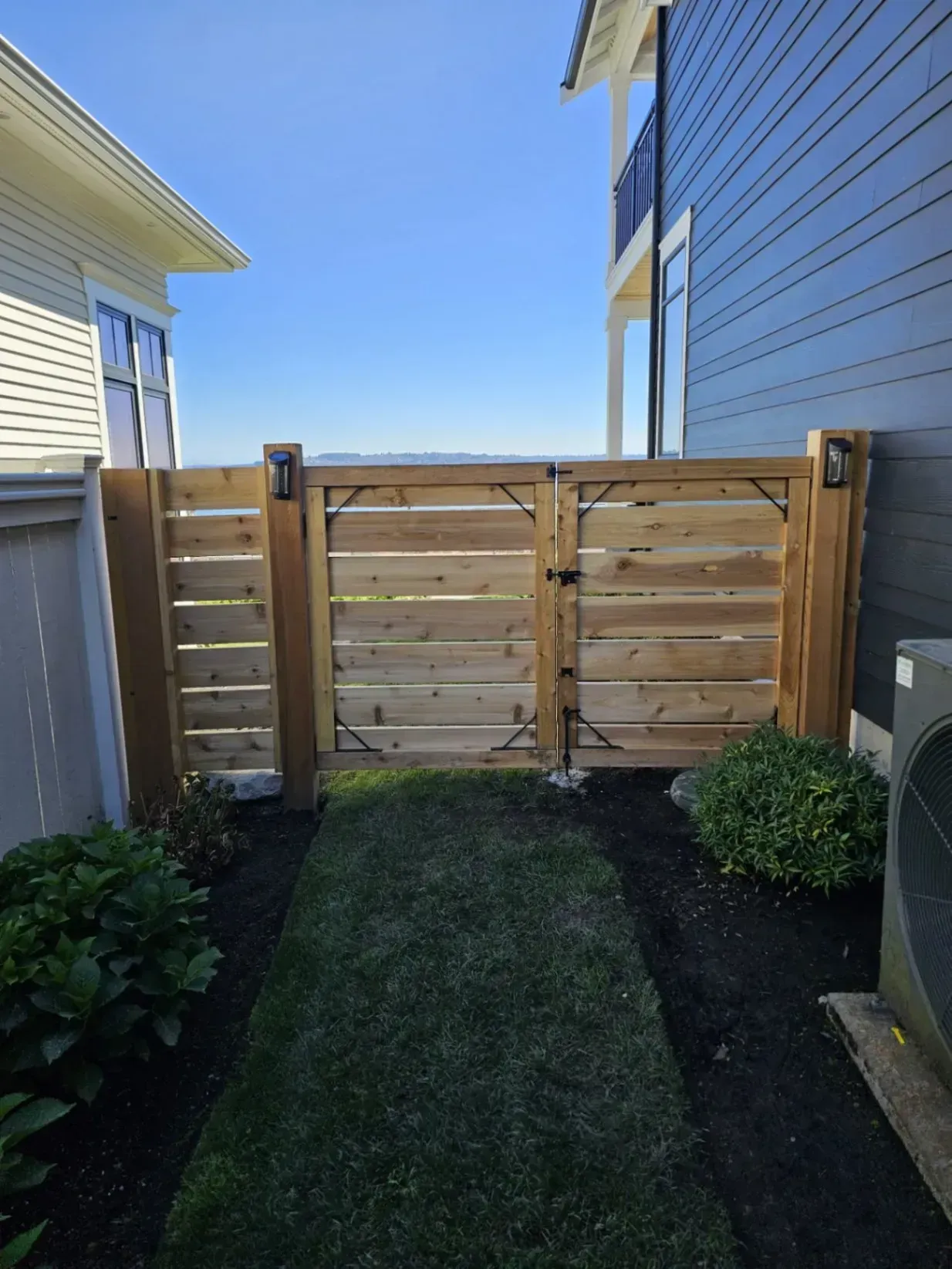 Wooden gate in a narrow pathway between two buildings, with plants and grass, on a sunny day.