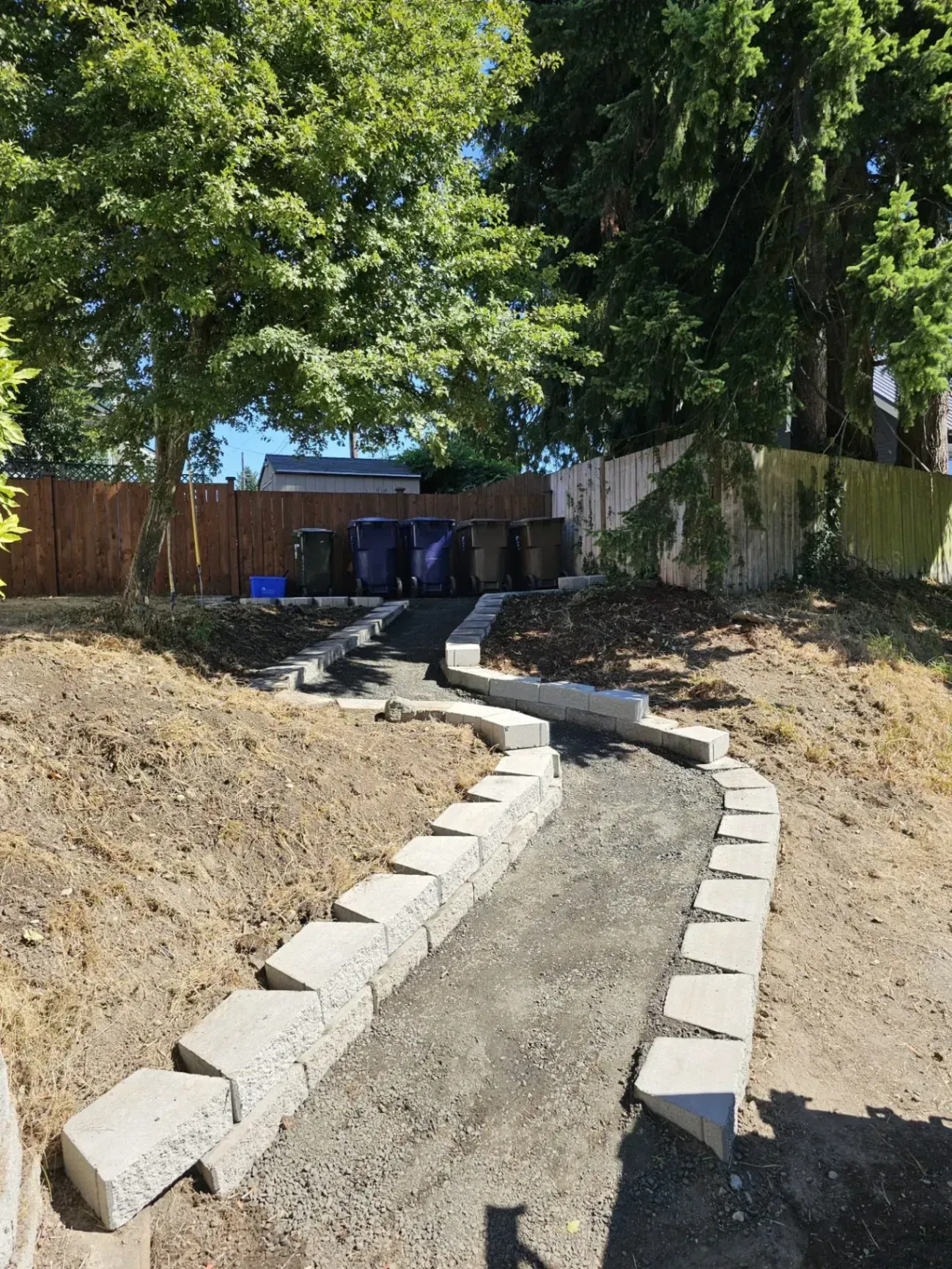 A gravel pathway with stone borders leads uphill to trash cans, trees in the background.