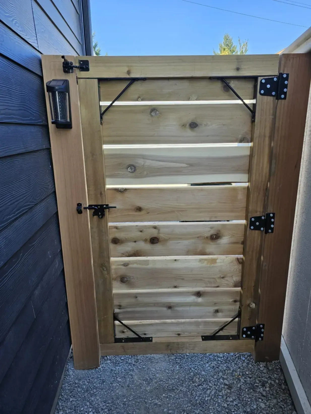 Wooden gate, horizontally slatted, with black hardware and solar light, between two walls.