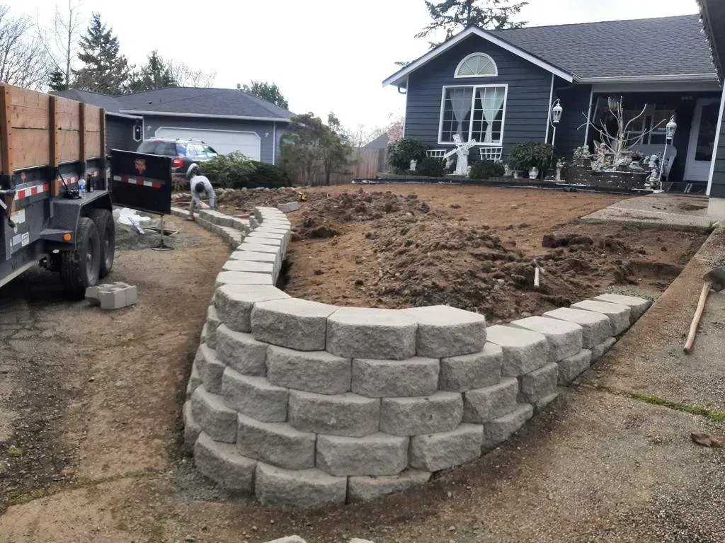 A gray stone retaining wall curves along a driveway in front of a house; a trailer is parked nearby.