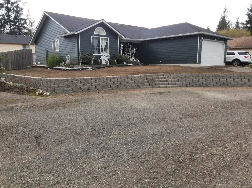 House with gray siding, a garage, and a retaining wall in front. Overcast sky.