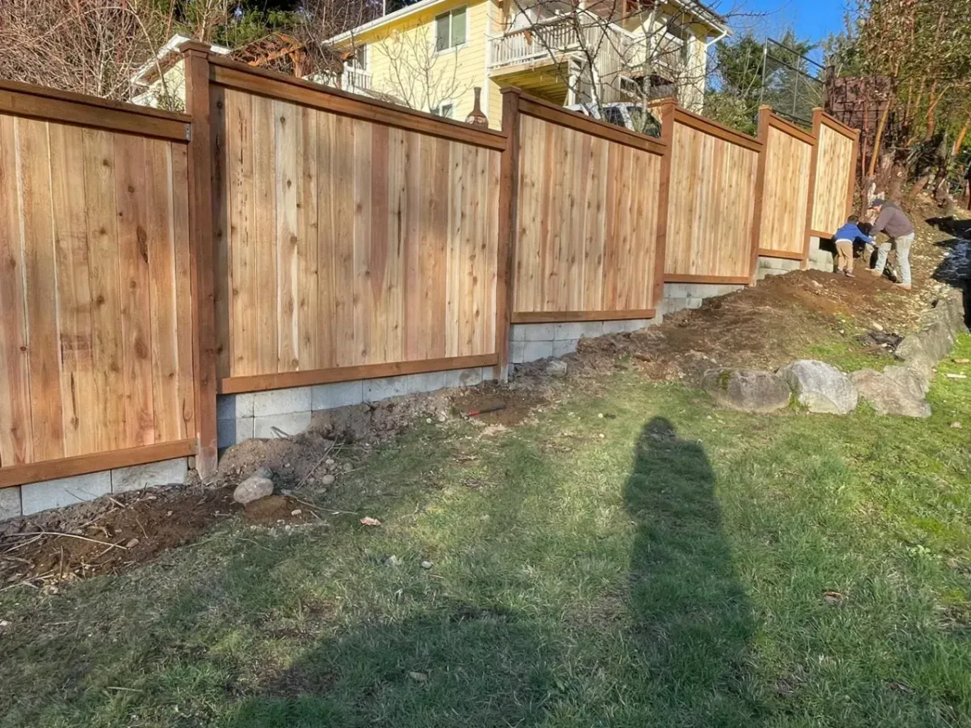 Wooden fence on a retaining wall, two people working on the right side, and green grass in front.
