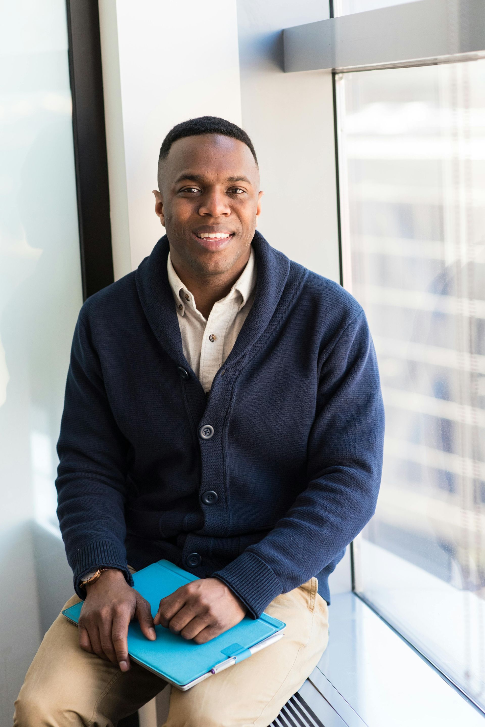 A man in a blue sweater is sitting on a window sill holding a tablet.