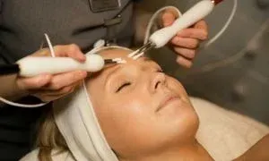 Woman receiving a facial treatment with handheld devices, eyes closed, in a spa setting.