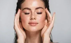 Woman with closed eyes, hands on temples, freckled face, peach-toned skin, wet hair, neutral background.