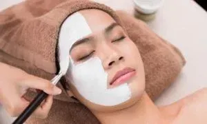 Woman receiving a facial, half her face covered in white mask, being applied with a brush, lying on a towel.
