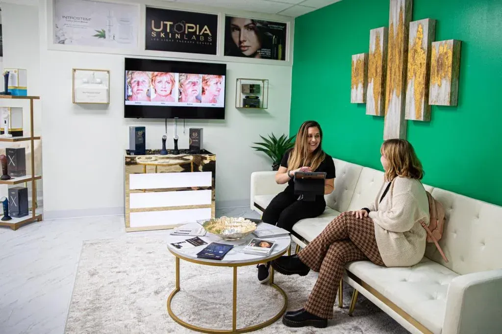 Two women in a modern waiting room; one discusses something while the other listens.