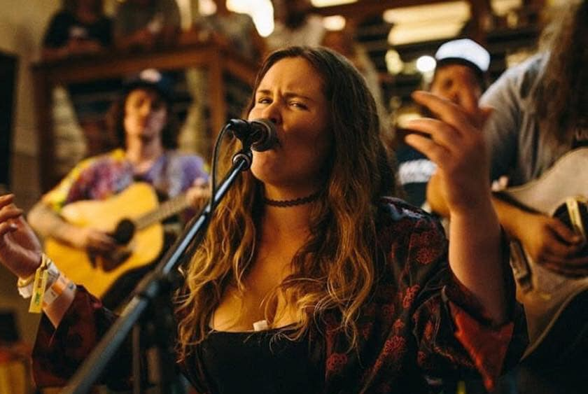 A woman is singing into a microphone while a man plays a guitar in the background.