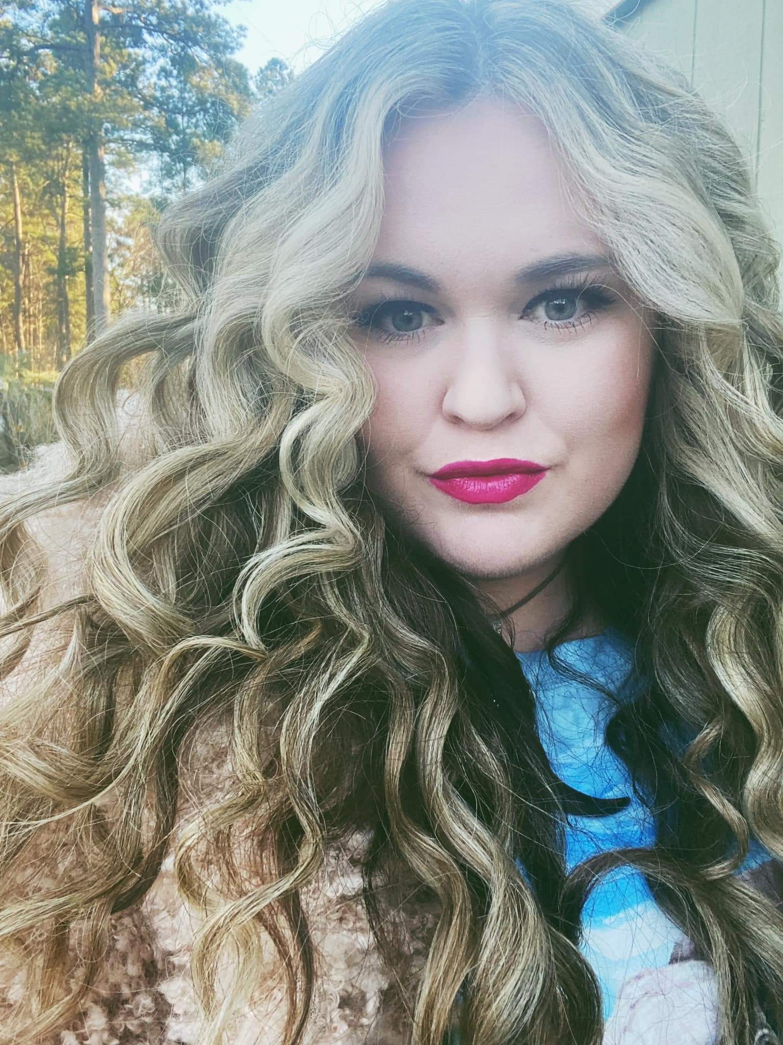 A woman with long curly hair and red lipstick is taking a selfie.