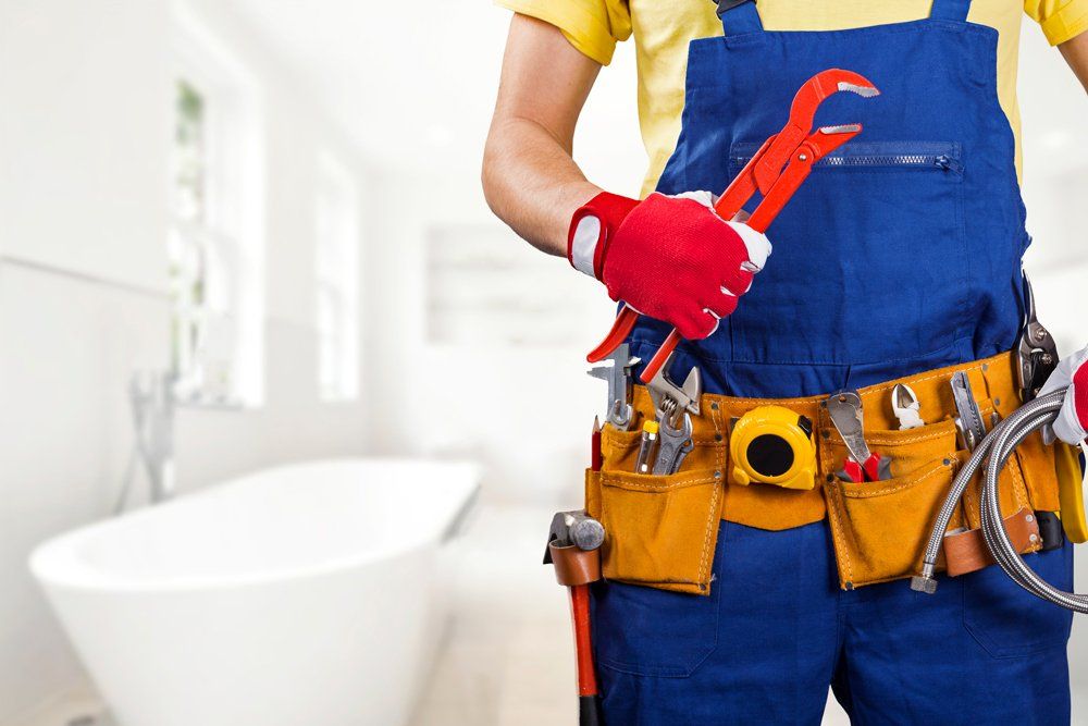 Plumber Doing Sink Pipe Repair — Albuquerque, NM — Donner Plumbing & Heating Plumber in blue overalls holding a red pipe wrench beside a bathtub in a bright bathroom