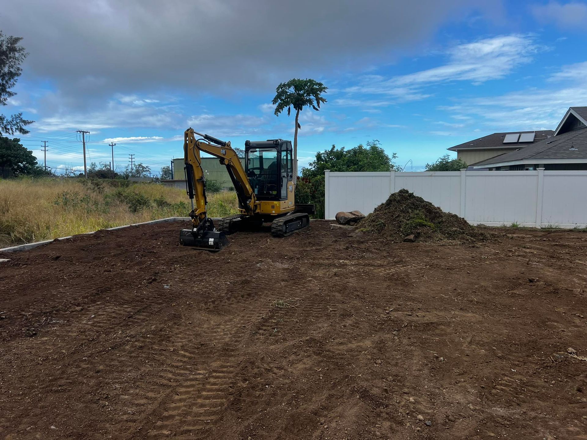 A yellow excavator parked on a cleared, dirt construction site next to a white fence under a blue sky.