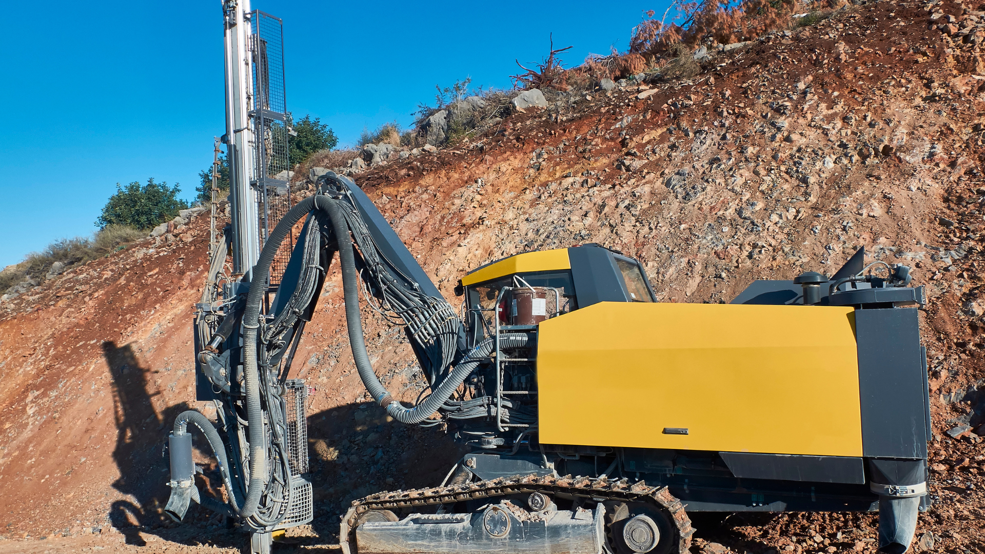 A yellow crawler drill rig operates on a steep, rocky dirt hillside under a clear blue sky.