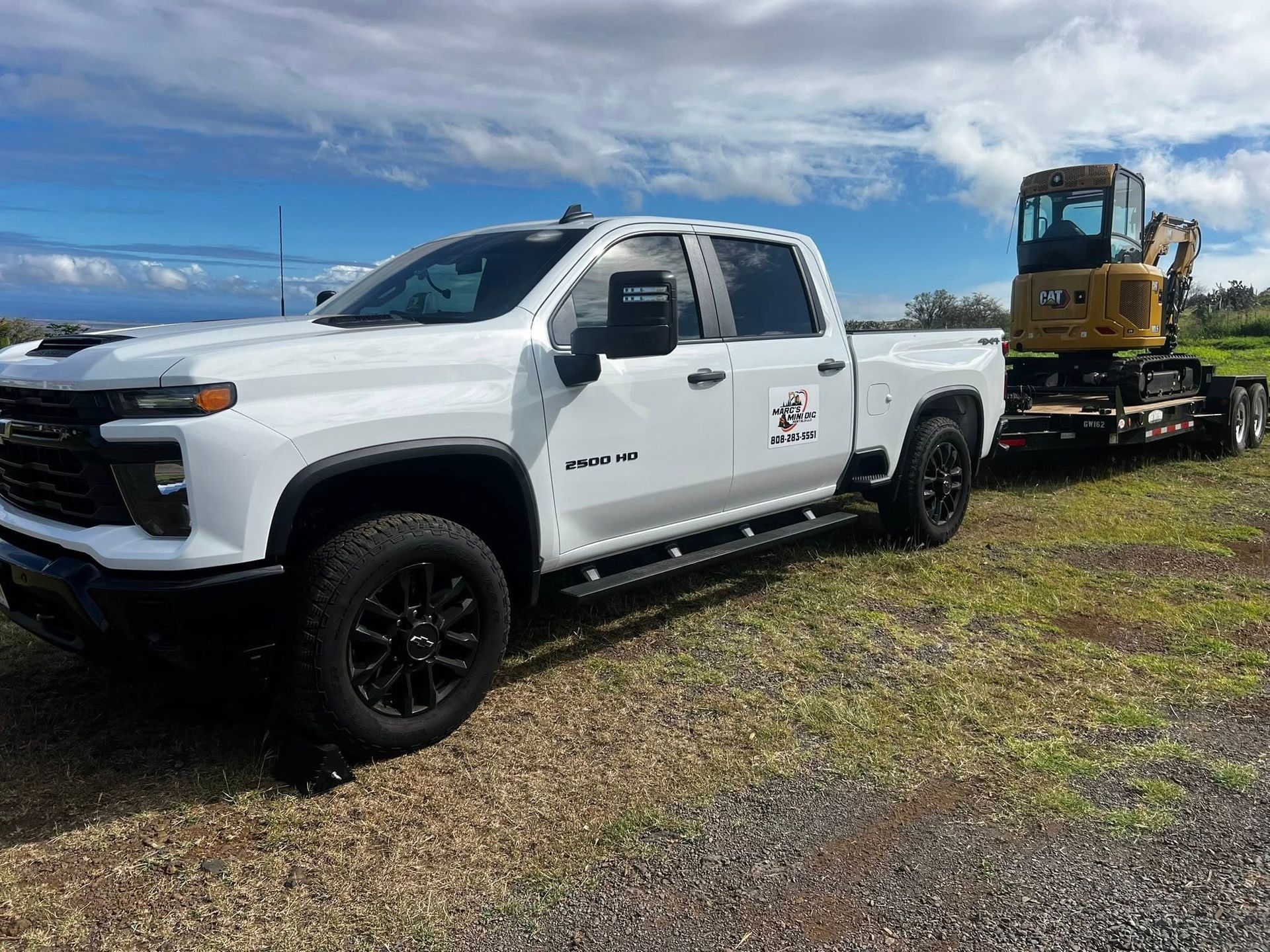 A white Chevrolet Silverado pickup truck hauling a yellow excavator on a flatbed trailer in a grassy field.