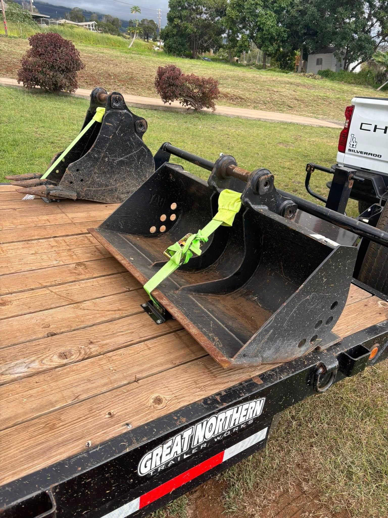 Two black excavator buckets secured with neon green tie-down straps on a flatbed trailer outdoors.
