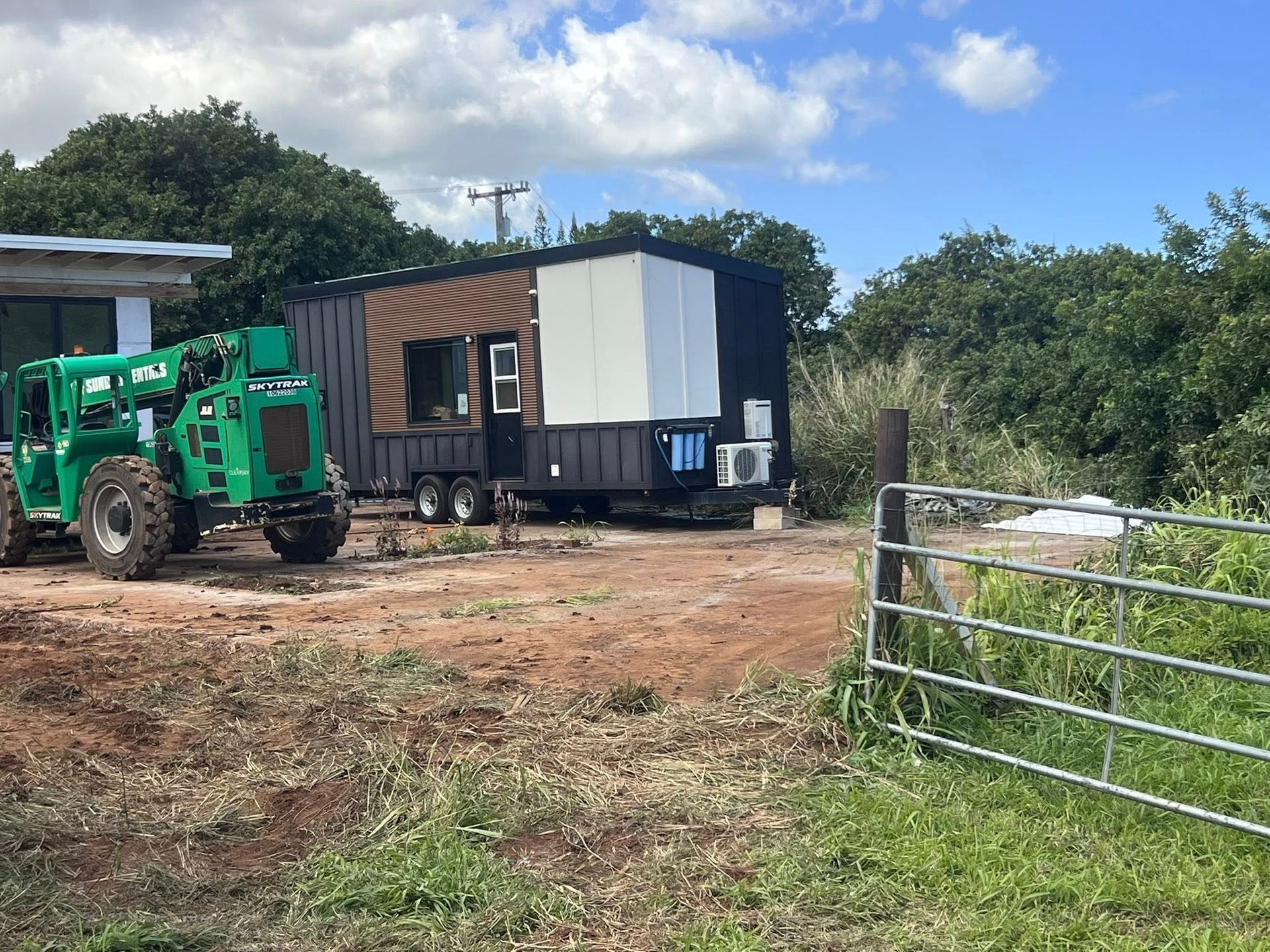 A green construction telehandler parked next to a modern, black and brown tiny home trailer on a rural, dirt lot.