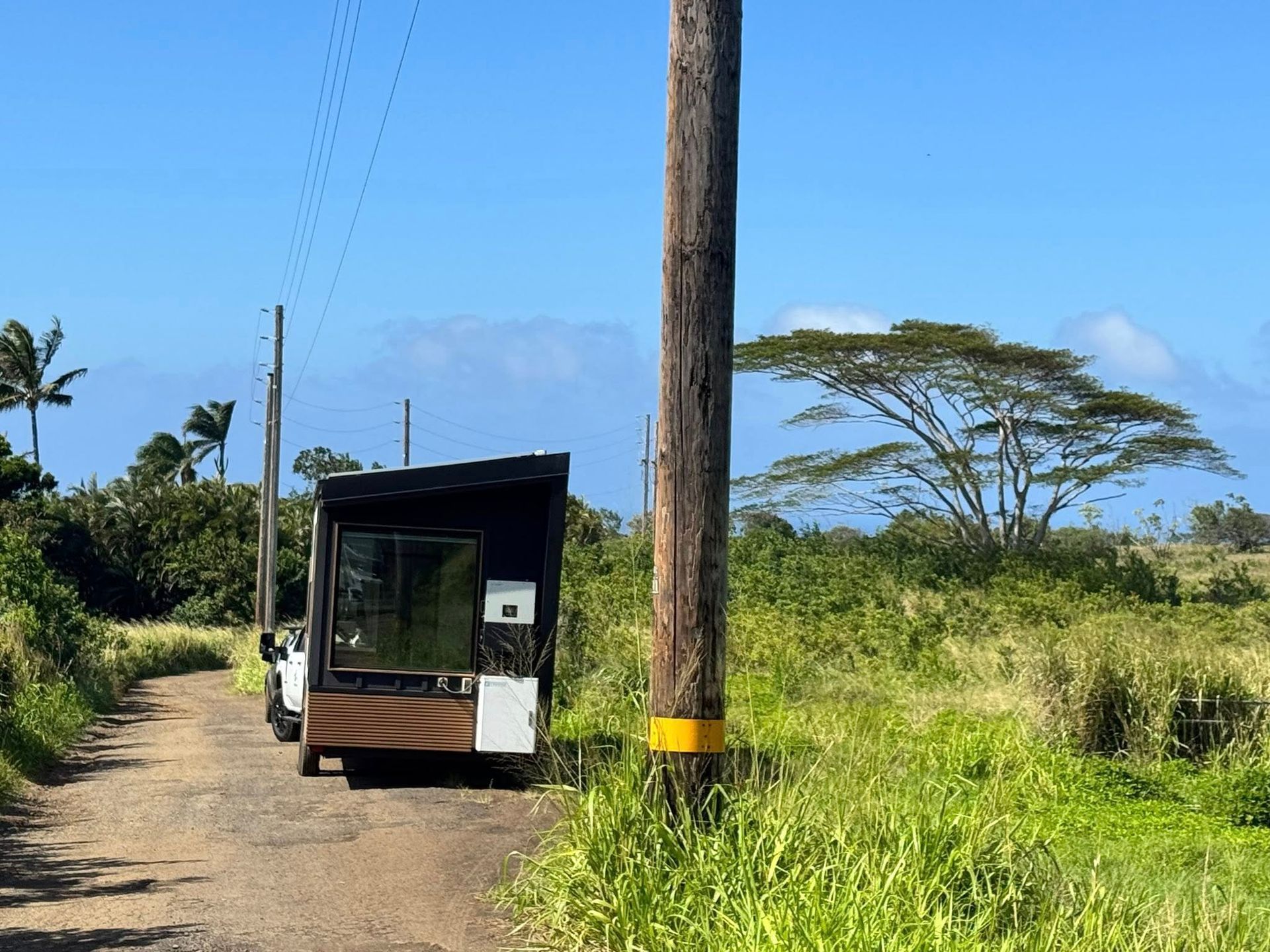 A modern, dark-sided tiny house parked on a dirt path next to a utility pole in a grassy, tropical field.
