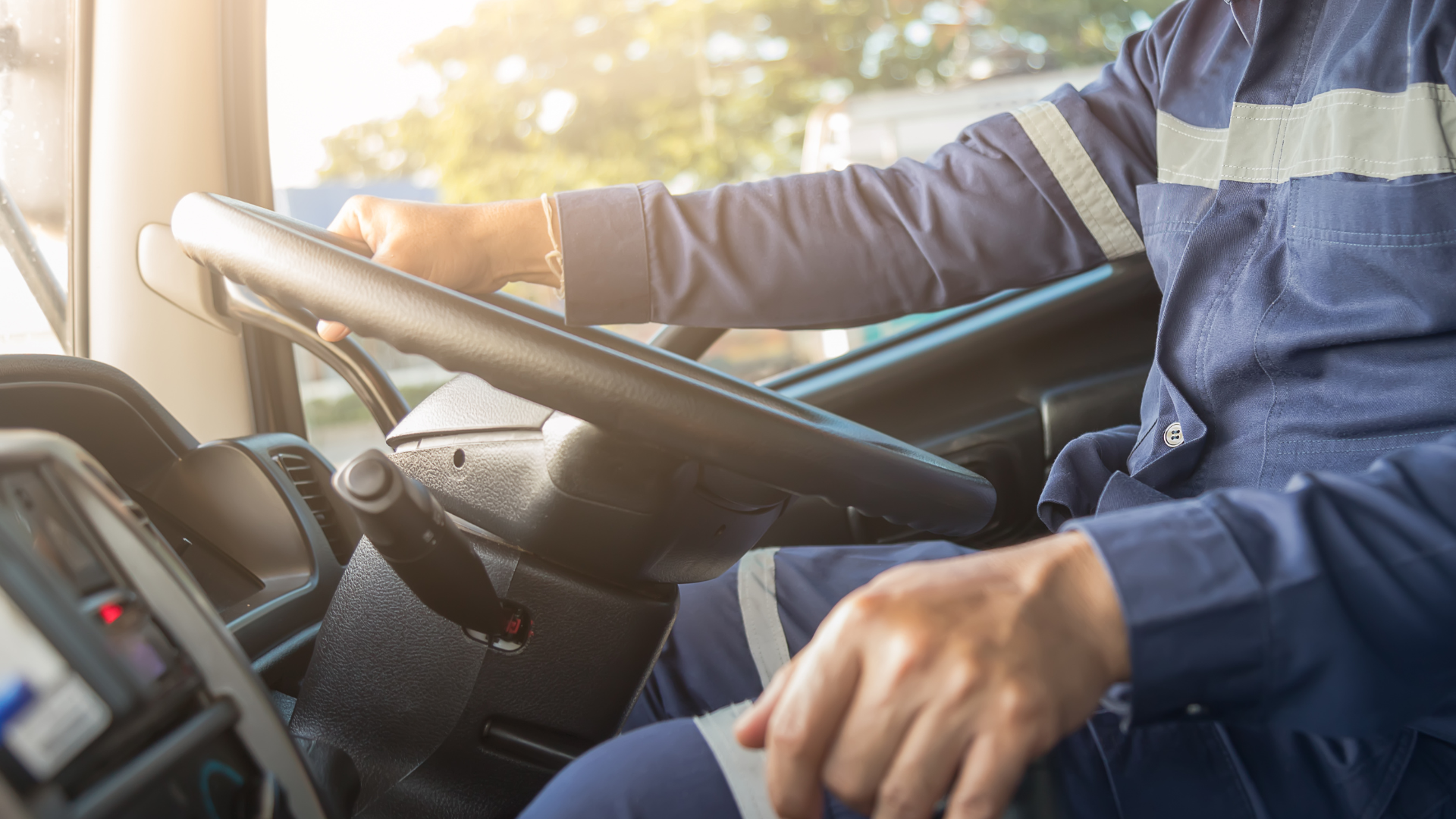 A person in a blue uniform with reflective strips driving a truck, hands on the steering wheel, in a bright, sunlit cab.