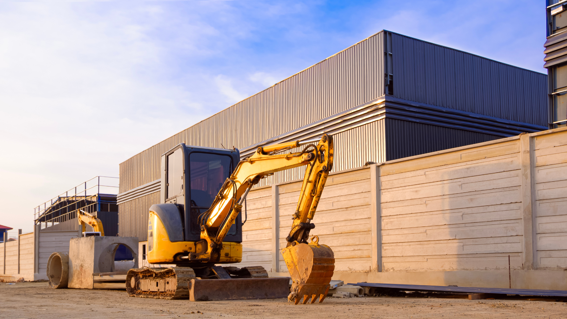 Two yellow excavators parked on a construction site next to a large metal building and a precast concrete wall.