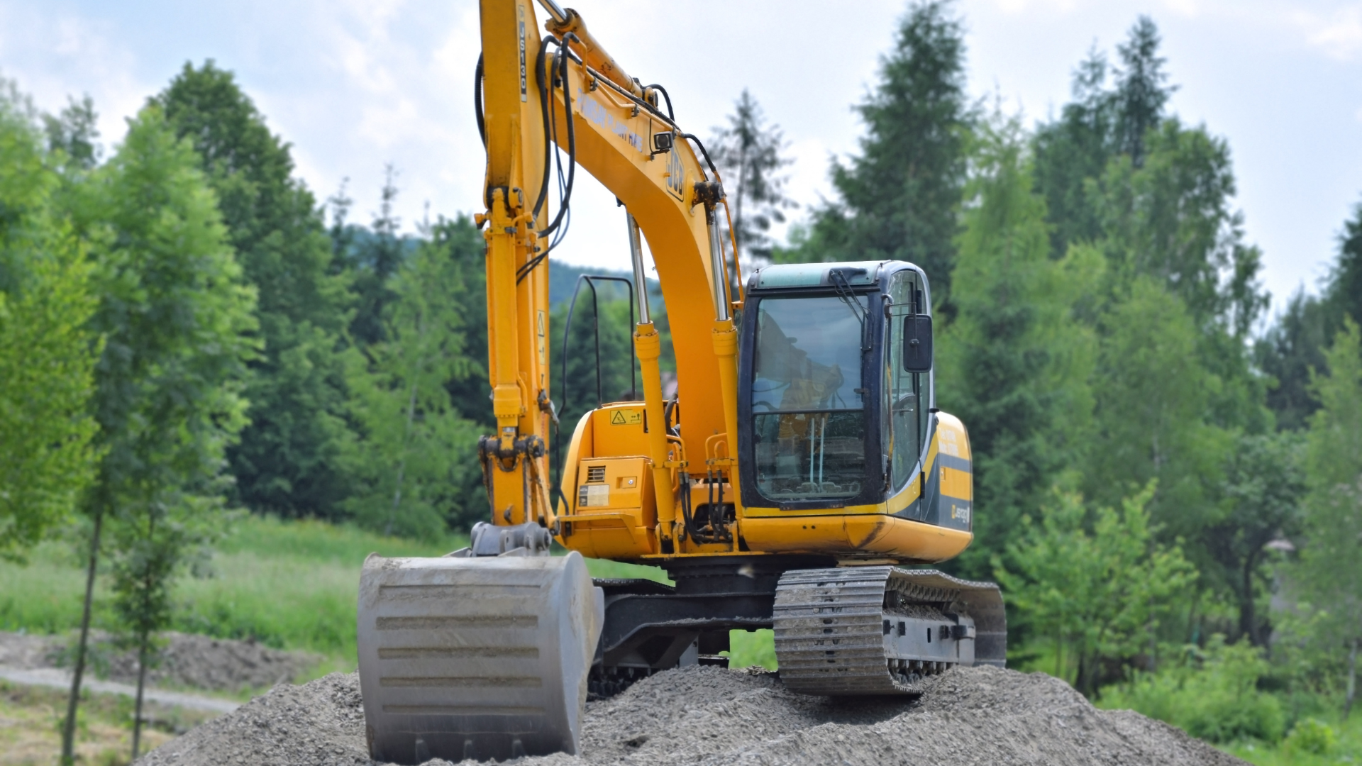 A yellow excavator sits atop a gravel pile in a lush, green wooded setting.