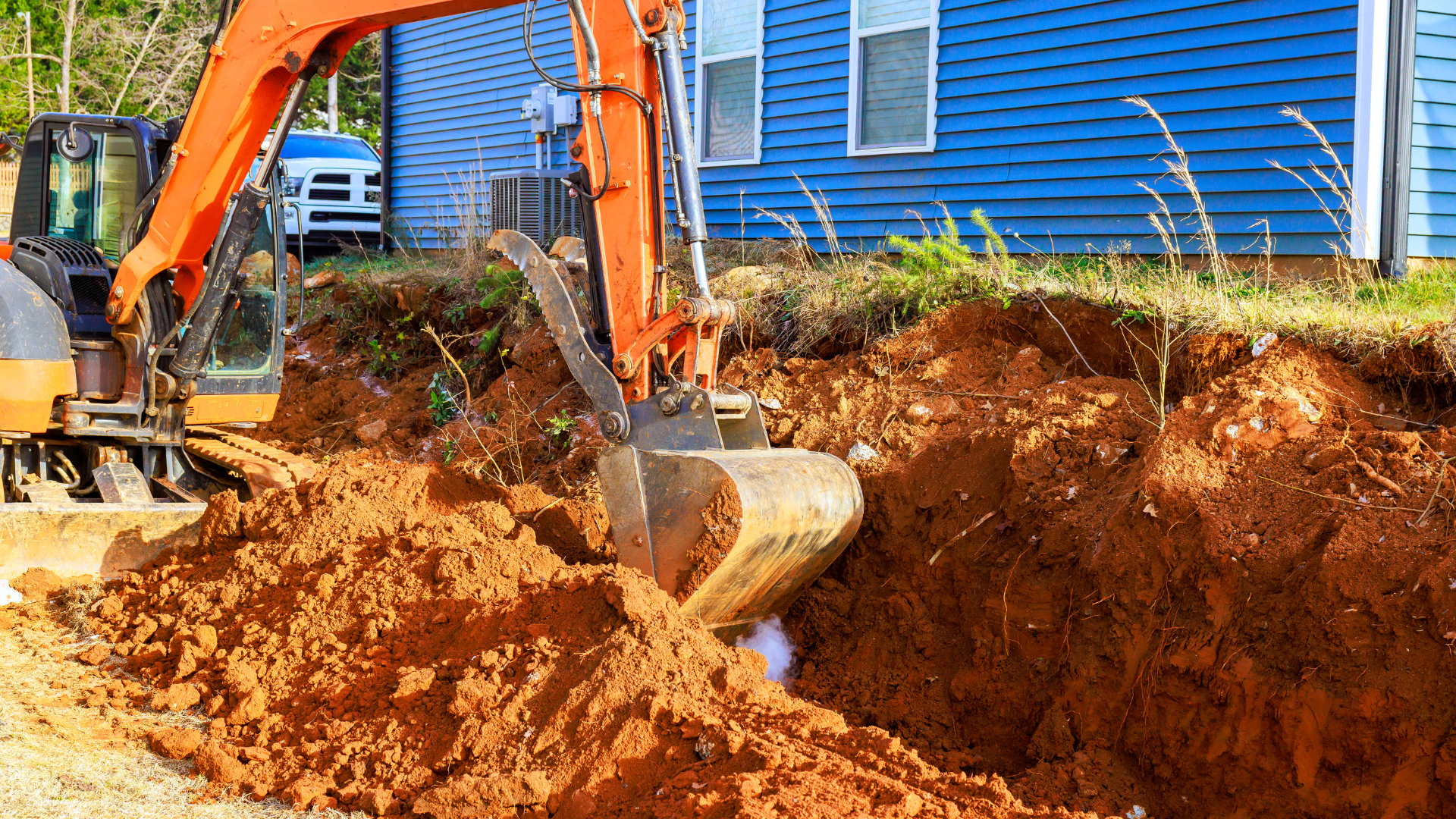An orange excavator digs a trench in red clay soil next to a blue-sided house.