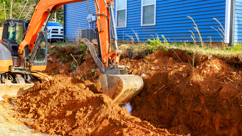 An orange excavator digs a trench in red clay soil next to a blue-sided house.
