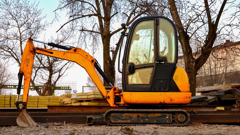 A yellow mini-excavator parked on a gravel construction site with trees and a building in the background.