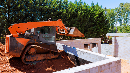 An orange skid steer loader operates near the brick foundation of a house under construction.