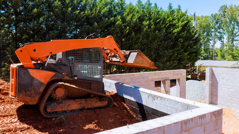 An orange skid steer loader operates near the brick foundation of a house under construction.