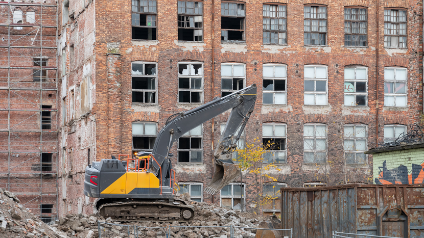 A yellow and grey excavator works amidst rubble in front of a derelict, multi-story brick building.